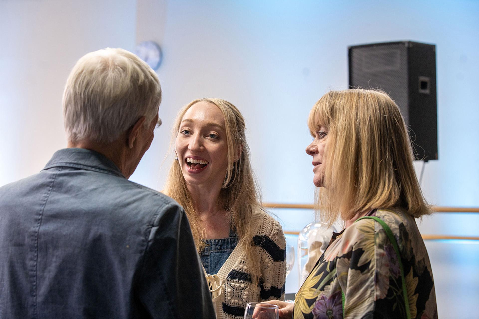 Smiling dancer with long blonde hair framing her face talks with a supporter with short grey hair, their back turned to the camera while another wearing a shirt with bright flowers looks on,