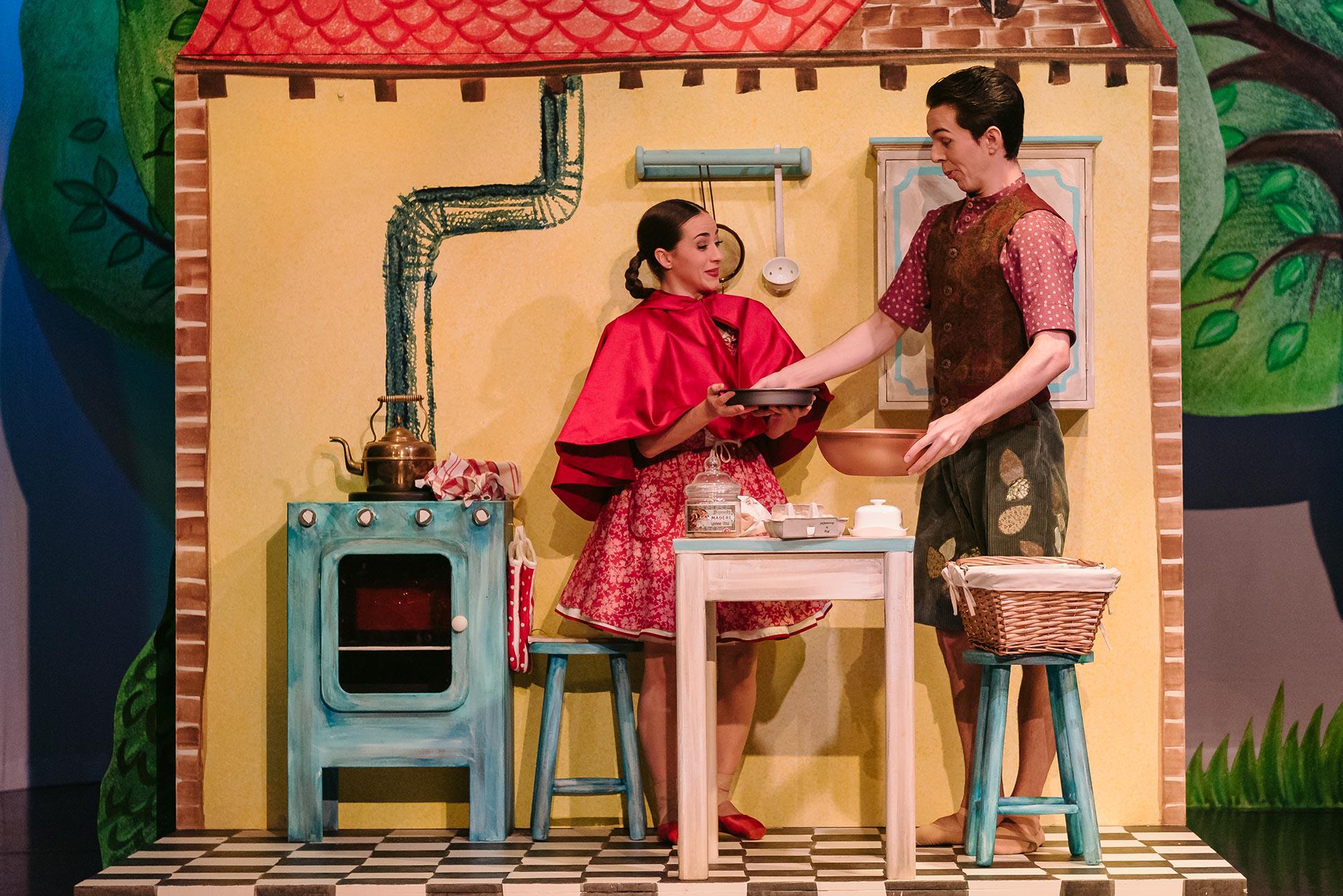 Two dancers stand on a set made to look like a yellow kitchen. One is wearing a red dress and the other a brown shirt. They are baking a cake.