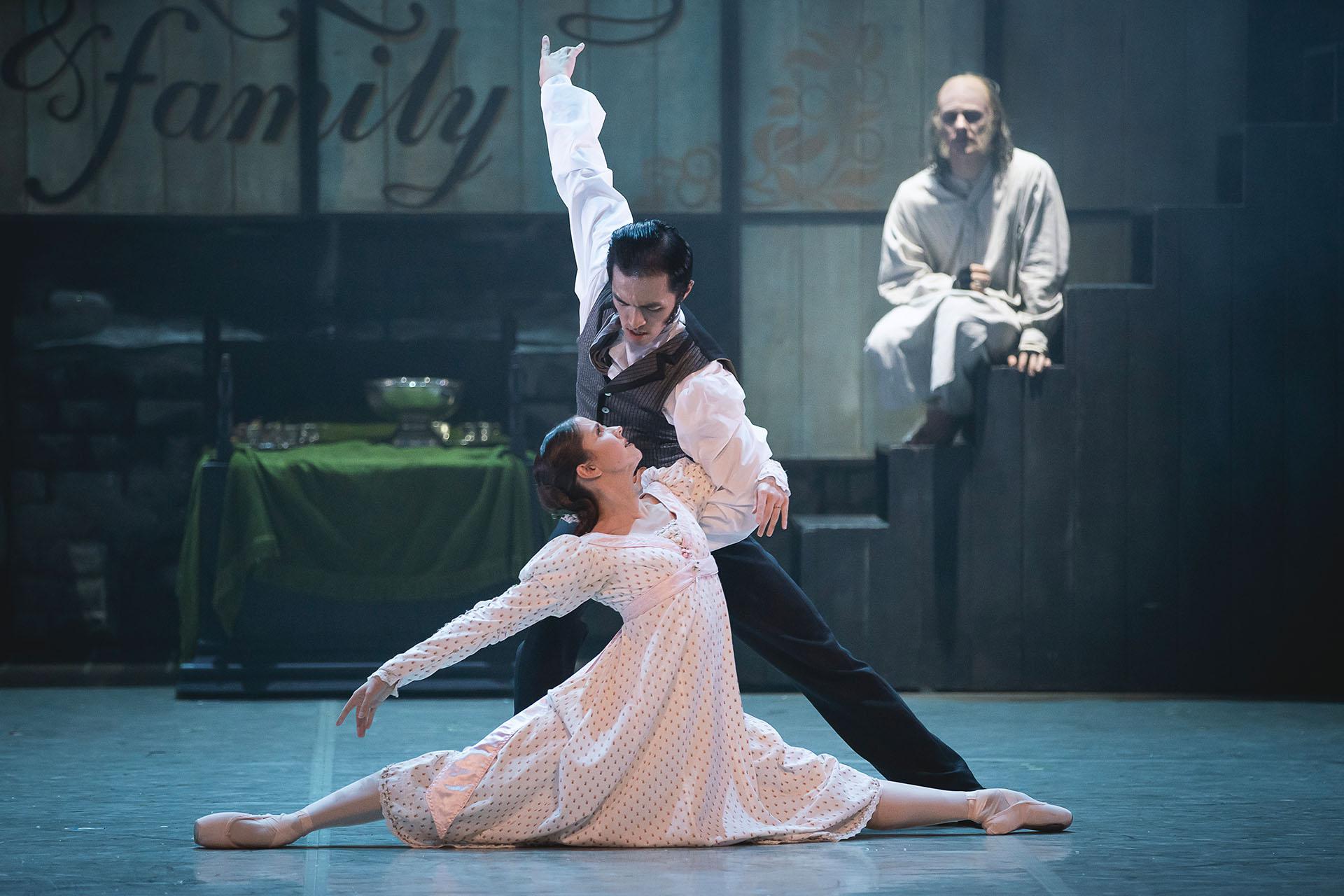 While Scrooge looks on, his younger self in white shirt and waistcoat dances with Belle who is wearing a white dress