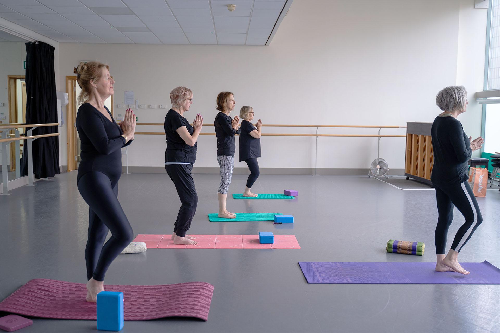 Five woman on brightly coloured yoga mats, hands together and standing on one foot