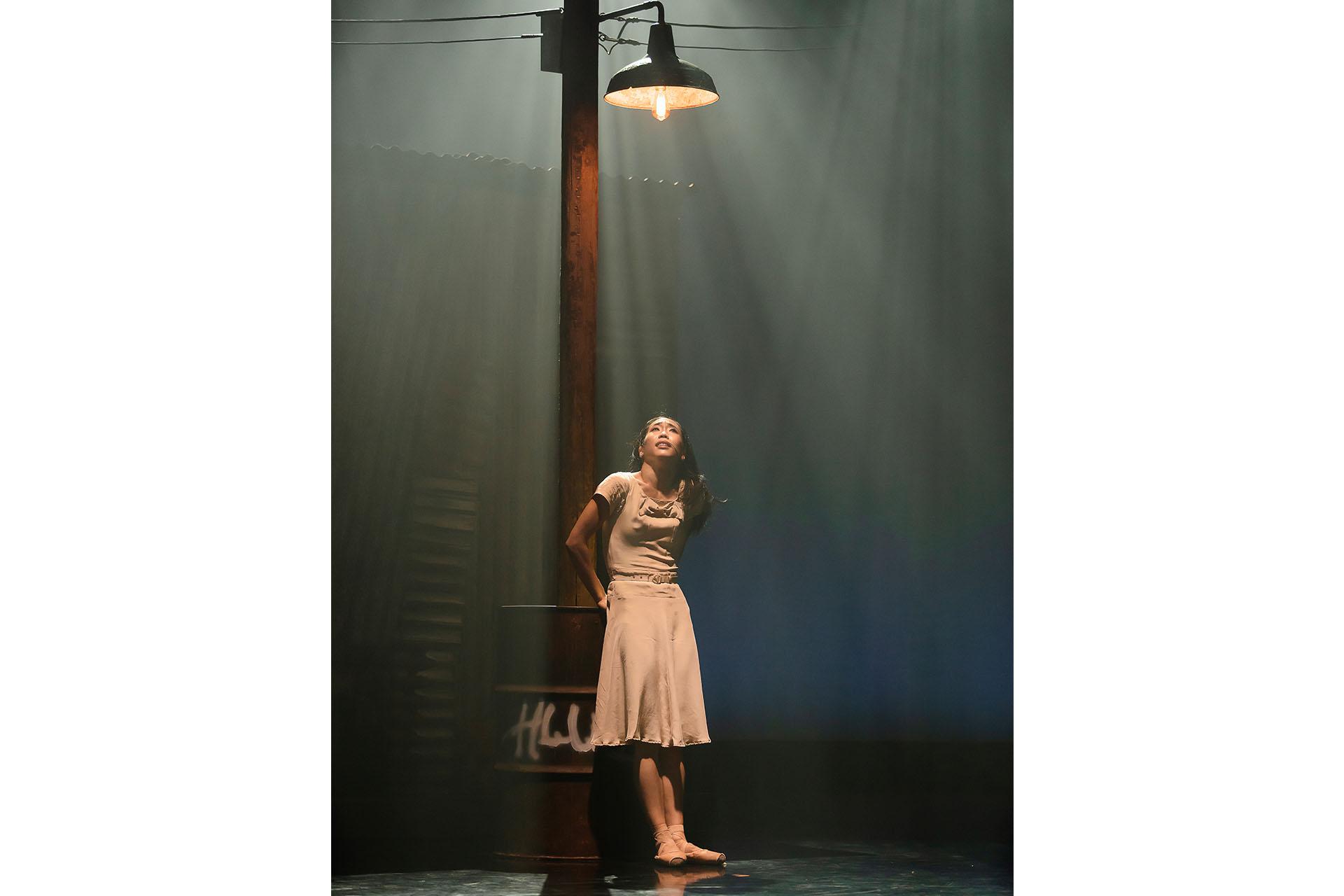 Woman with dark hair tied back stands under a streetlight in the gloom