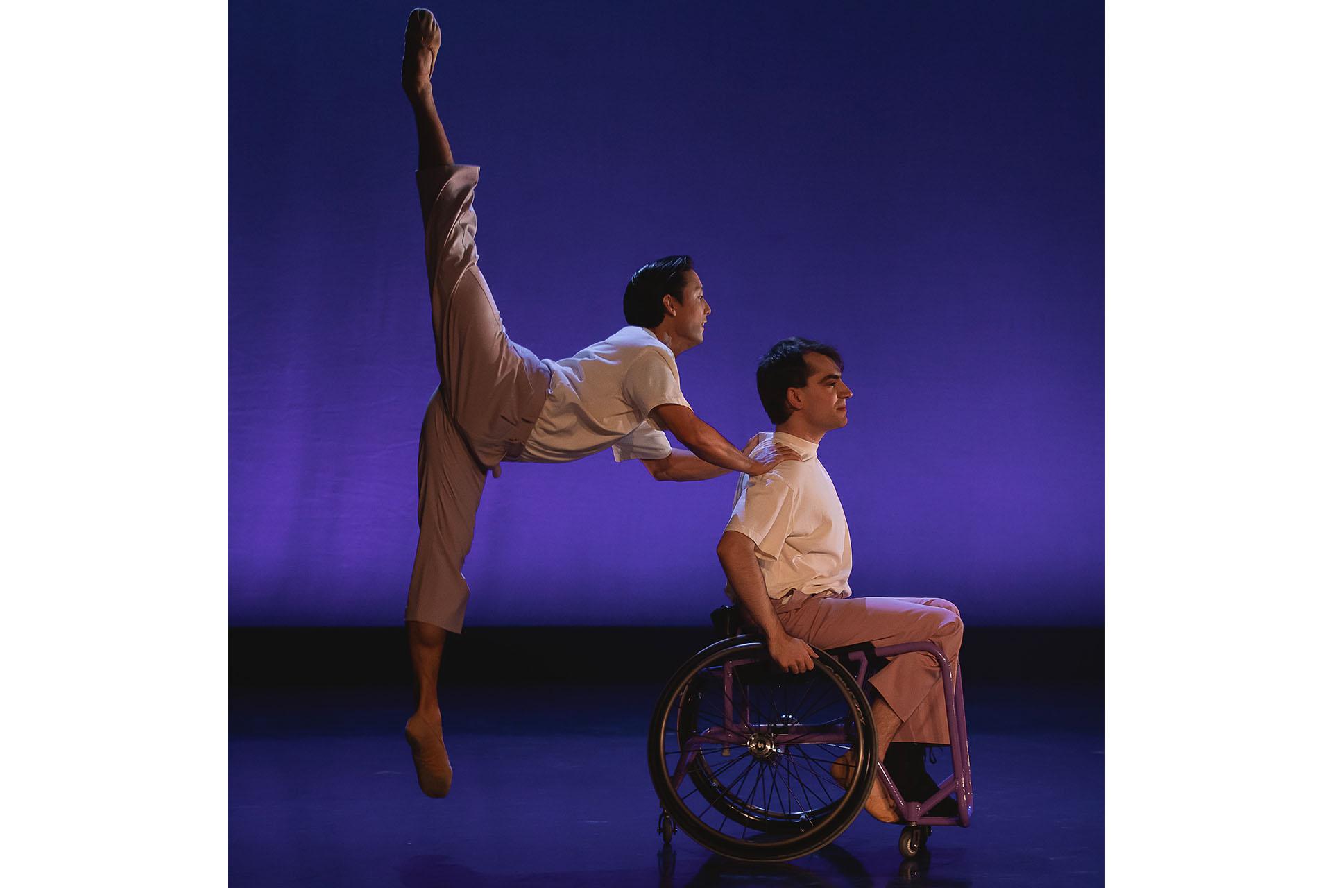 Dancer in a wheelchair in profile with another dancer, hands on his shoulders jumping with one leg pointed down and the other pointed up