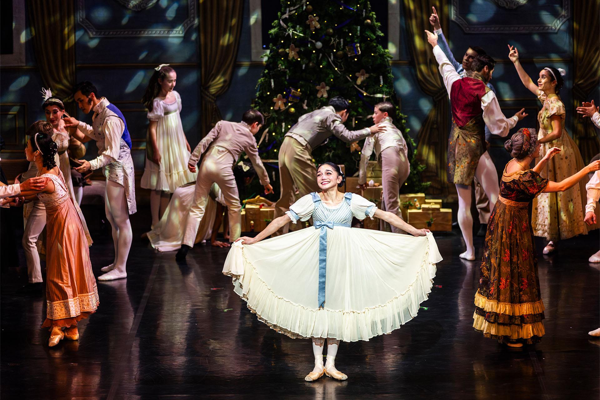 Girl holds the hem of her blue trimmed white skirt and smiles broadly as the family and party guests bustle around her