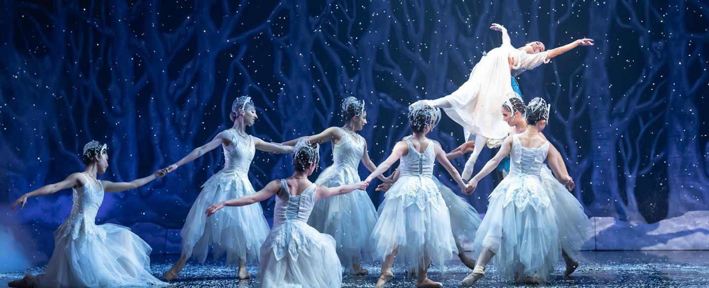 Dancers dressed in white in a whiter scene hold a young, happy woman aloft