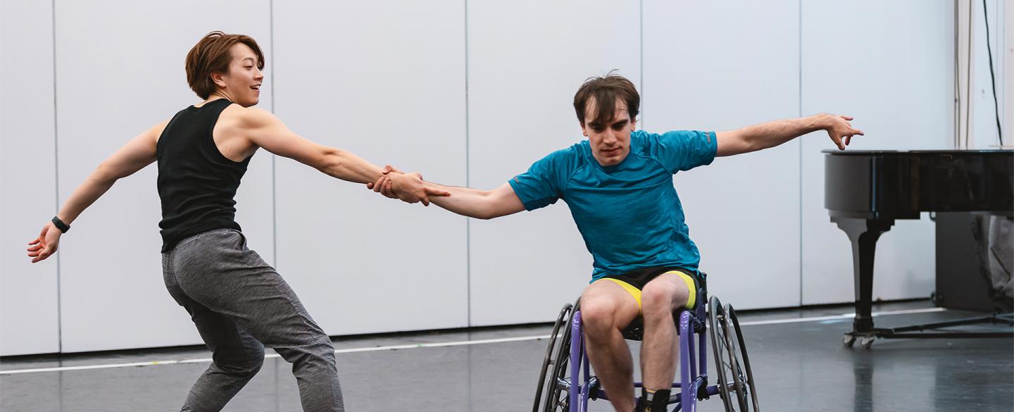 Two dancers hold hands and pull away from each other, one braced with his feet on the ground, the other with the wheels of his chair against the pull