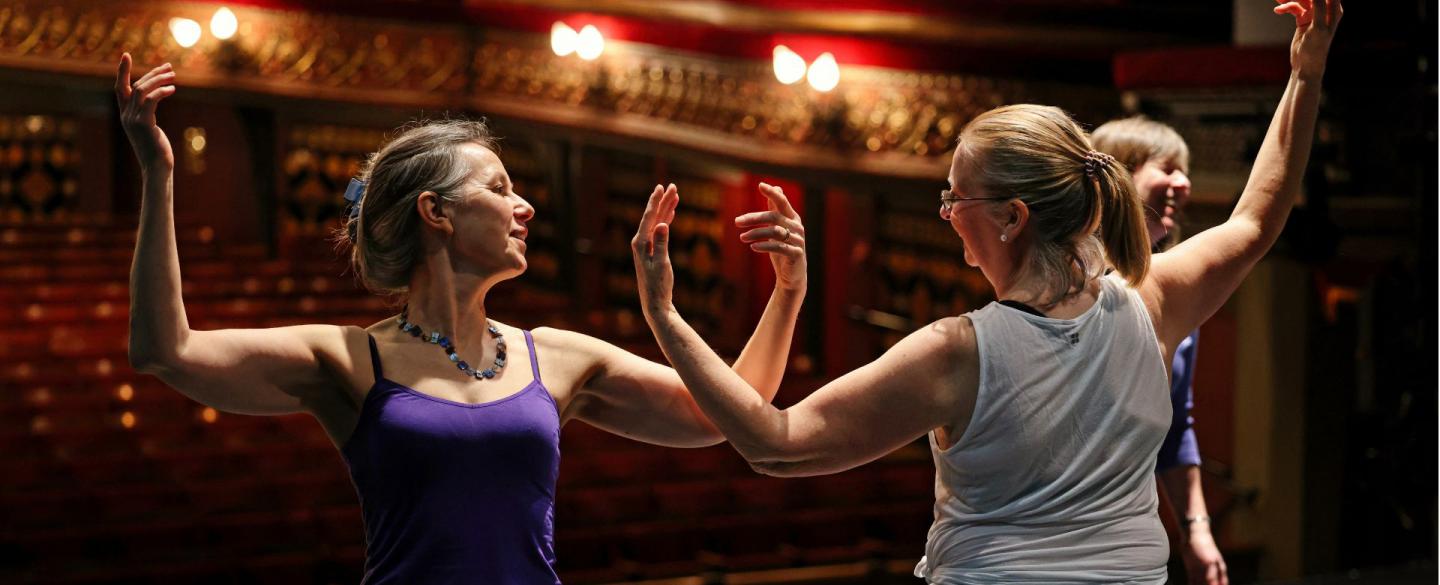 Two older dancers rehearse moves on a stage in front of an empty audience space. They stand side-by-side facing opposite directions. Their raise both their arms, nearly touching elbows whilst looking at each other.