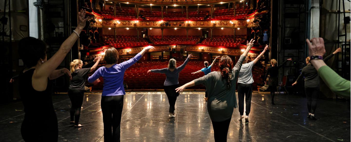 A group of older dancers on a stage stand separately and rehearse dance moves towards an empty multi-tiered audience space