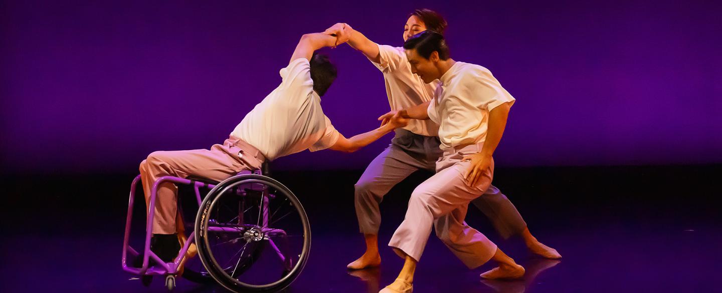 One dancer in a wheelchair leans back and holds the arms of two more dancers immediately behind him