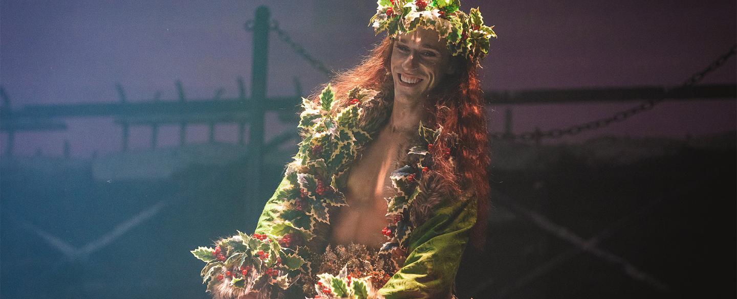 A dancer smiling on stage in a large green and red robe covered in holly with a holly crown