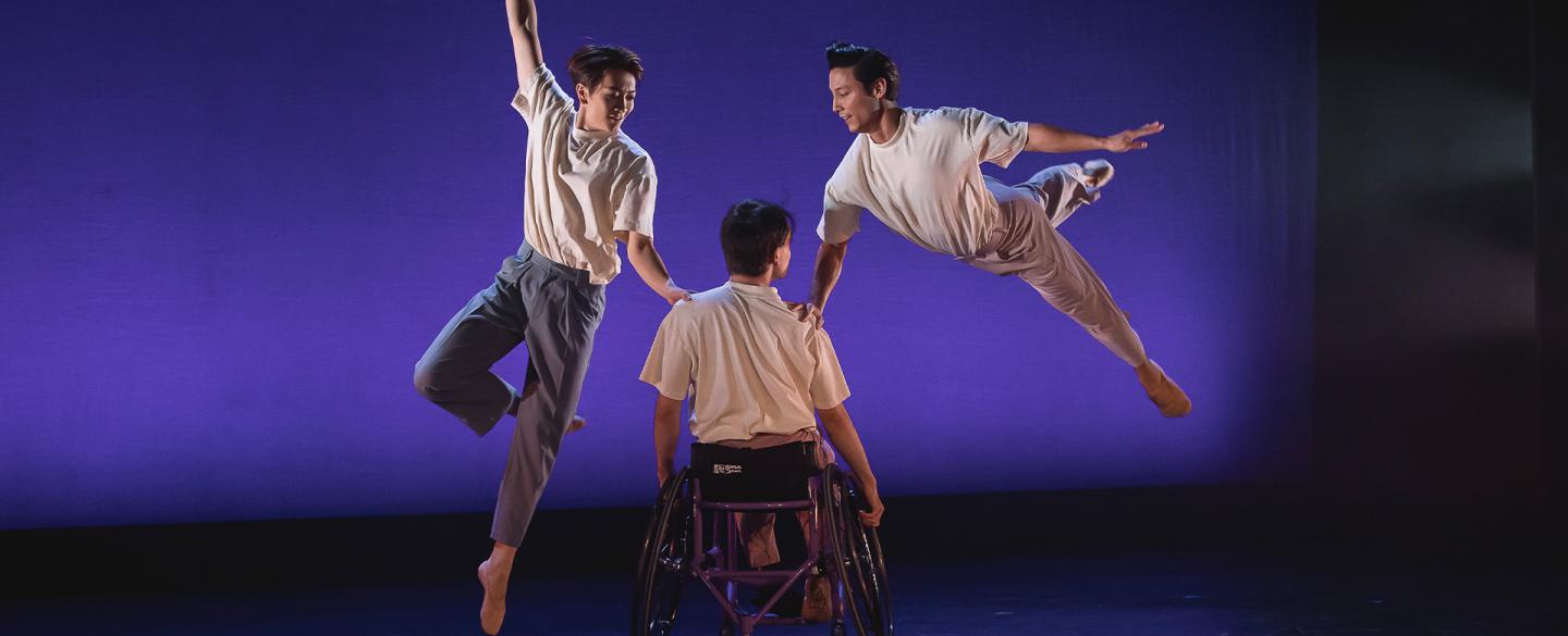 Three dancers on a purple stage, two dancers are leaping beside a dancer in a wheelchair