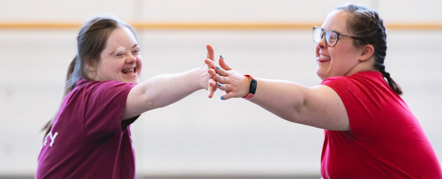 Two dancers in an Ability class sit across from each other on the floor, touching hands and smiling