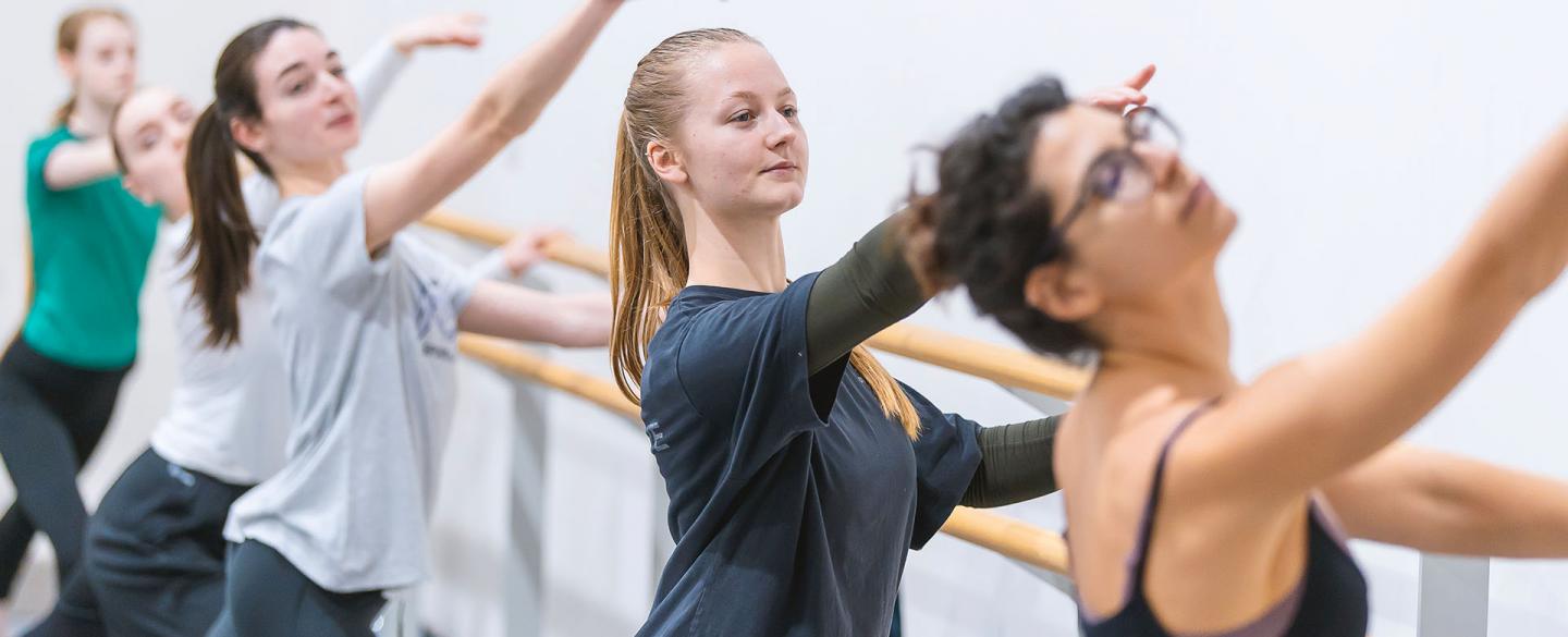 Students at the barre, holding on with one hand, their other raise deliberately above their head