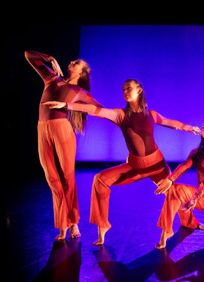 Three dancers bathed in red light against a background of black and blue, each in a different pose - one on her toes sood tall, the next bending one leg at 90 degrees, the third lower still