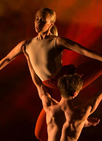 Bathed in red light and male dancer holds upa female dancer as she holds a pose with her arms straight and her fingers pointed down
