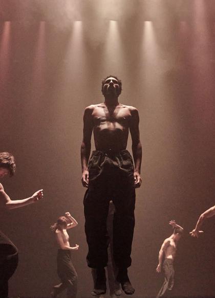 Five dancers in hazed light striking firm poses, one central and lower half of his face cast in shadow