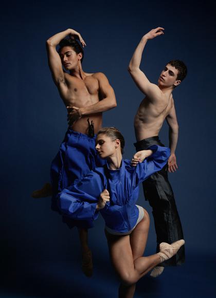 Three dancers against a blue backdrop. Two each have one arm raised above their heads