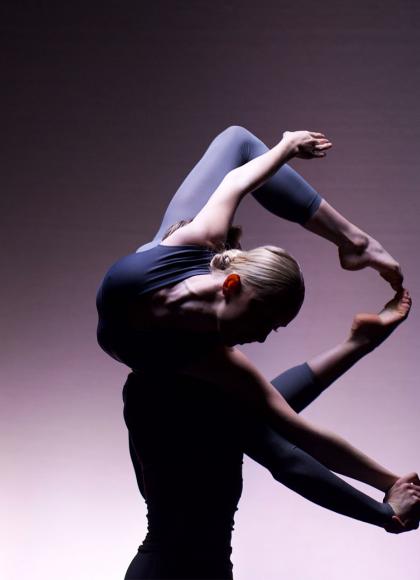 Against a gradiated purple backdrop one dancer lifts another, the lifted dancer horizontal, toes together but knees apart back curled around the lifters head