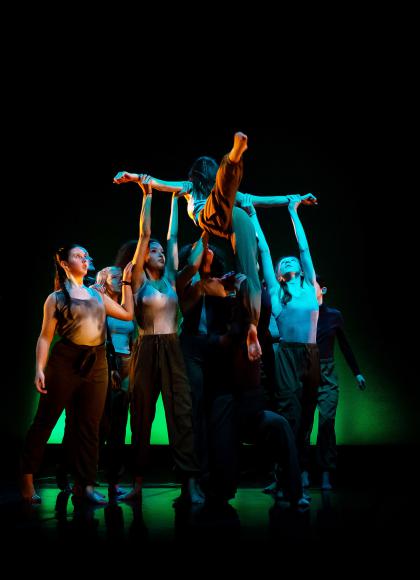 A dancer with leg outstretched in a low atmospheric light being lifted by a group of dancers underneath. 