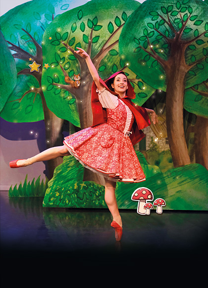 Dancer in red with a red hood and red pointe shoes stands arabesque smiling as she walks through a forest