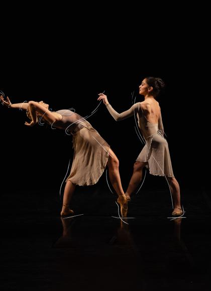 Against a black backdrop two dancers stand facing each other with one foot en pointe, and one dancer leaning back with her arms above her head