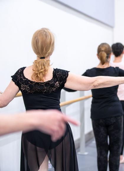 A row of dancers at the barre, one hand held out level with their shoulders, the other hand holding the barre, their ankles together in first