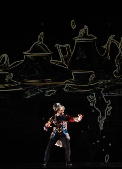 A man in a large top hat motions in front of giant, stylised crockery