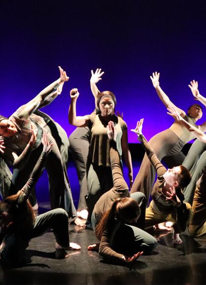 Against a blue and purple backdrop, a figure stands with one arm raised in a defiant gesture surrounded by dancers shielding themselves from her brilliance.