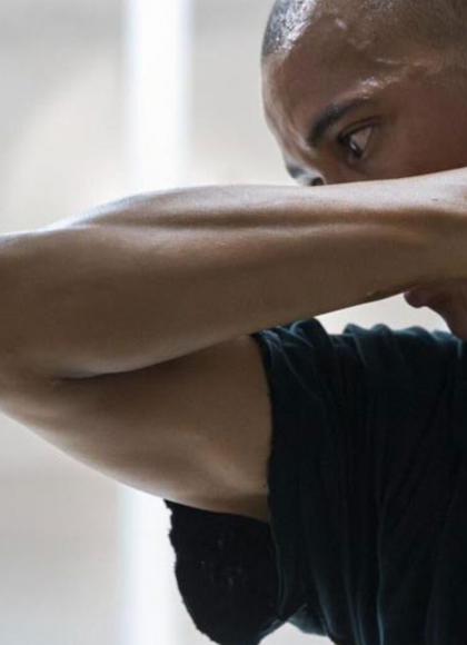 A dancer in a black shirt covers their face with their forearm while holding their hand to the side of their neck.