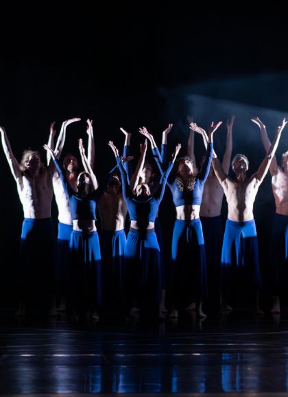 On a dark background, a group of dancers in blue outfits stand in a spotlight while reaching their arms up.