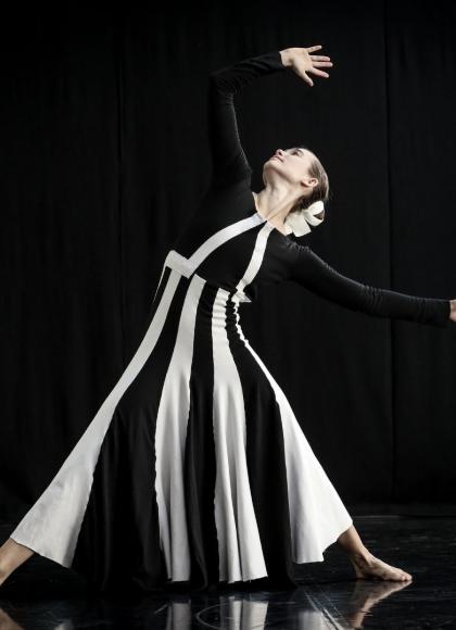 On a black background, a dancer in a black and white striped dress poses with an arm held above their head and their legs spread in a wide stance.
