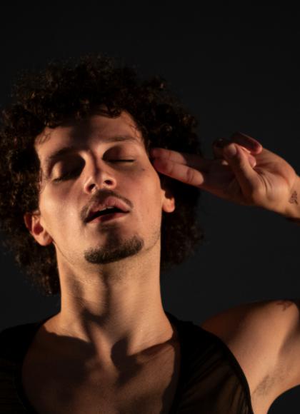 On a black background, a dancer faces the camera with their eyes closed and holds two fingers to the side of their head.