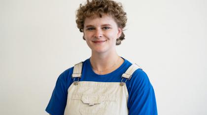 Martha is wearing a blue T-shirt, white dungarees, has short brown curly hair and is smiling straight at the camera