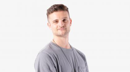 Man wearing a grey T-shirt, short brown hair, smiling wanly at the camera