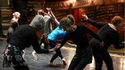 Older dancers rehearse ballet moves on a stage. Two stand in front of one another, bend forward and begin to lift their arm towards the other