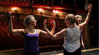 Two older dancers rehearse moves on a stage in front of an empty audience space. They stand side-by-side facing opposite directions. Their raise both their arms, nearly touching elbows whilst looking at each other.