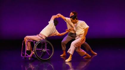 One dancer in a wheelchair leans back and holds the arms of two more dancers immediately behind him