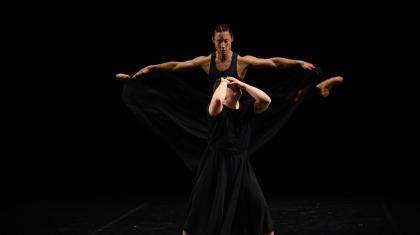 A dancer holds his face as another dancer leaps behind him with outstretched legs, looking down on the other dancer.