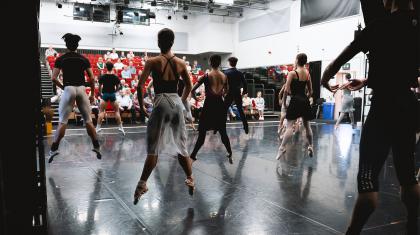 Dancers on stage in Company class, each in first jumping inches off the ground being watched by supporters