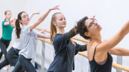 Students at the barre, holding on with one hand, their other raise deliberately above their head