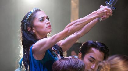 Wielding a dagger with outstretched arms and looking pained, Ygraine wears a blue dress, her arms muscular