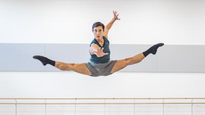 A dark haired dancer leaps, legs spread mid-jump, one hand outstretched to the camara