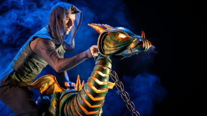 A young dragon, his eyes glowing and light visible through his scales; chained tied to his neck and a puppeteer posing him dramatically.