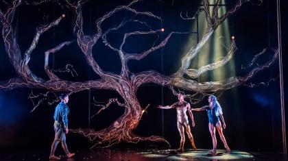 Under a leafless tree, Merlin approaches the beckoning Lady of the Lake wearing blue and Helios in golden reflective clothes.