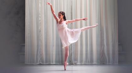 A dancer in a white dress stands on pointe with arms and legs outstretched