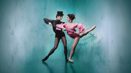 Two women, one in black with black pointe shoes and hat supporting another woman with a walking stick, her wearing pink