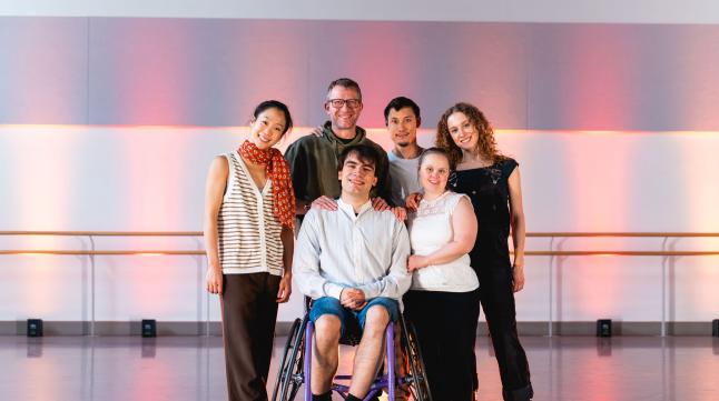 Six people stand in front of warm pink smiling and facing the camera